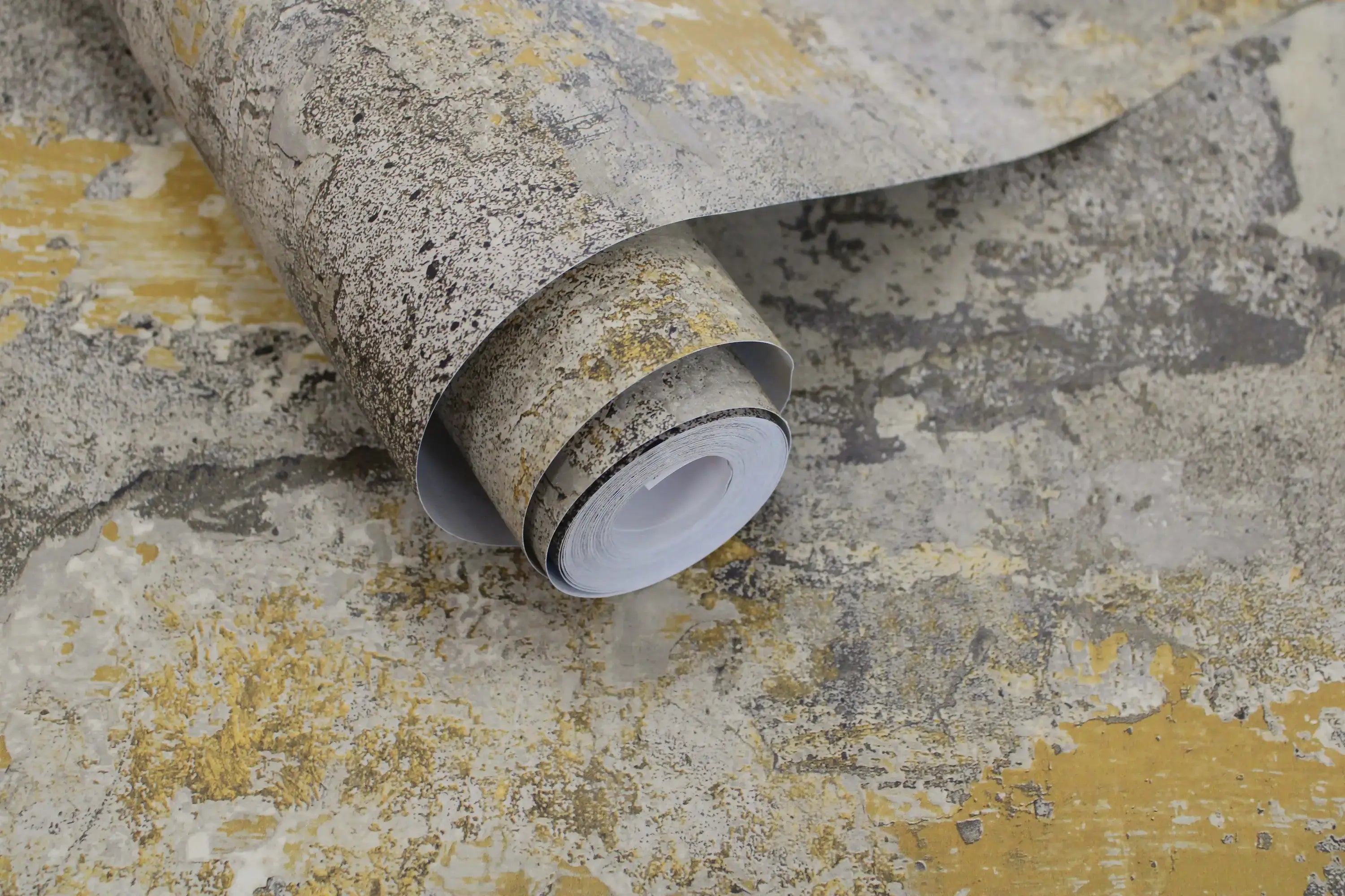 En rulle af Elgon - Ochre tapet vist ovenpå tapetets samme design, der fremhæver de rustikke detaljer.