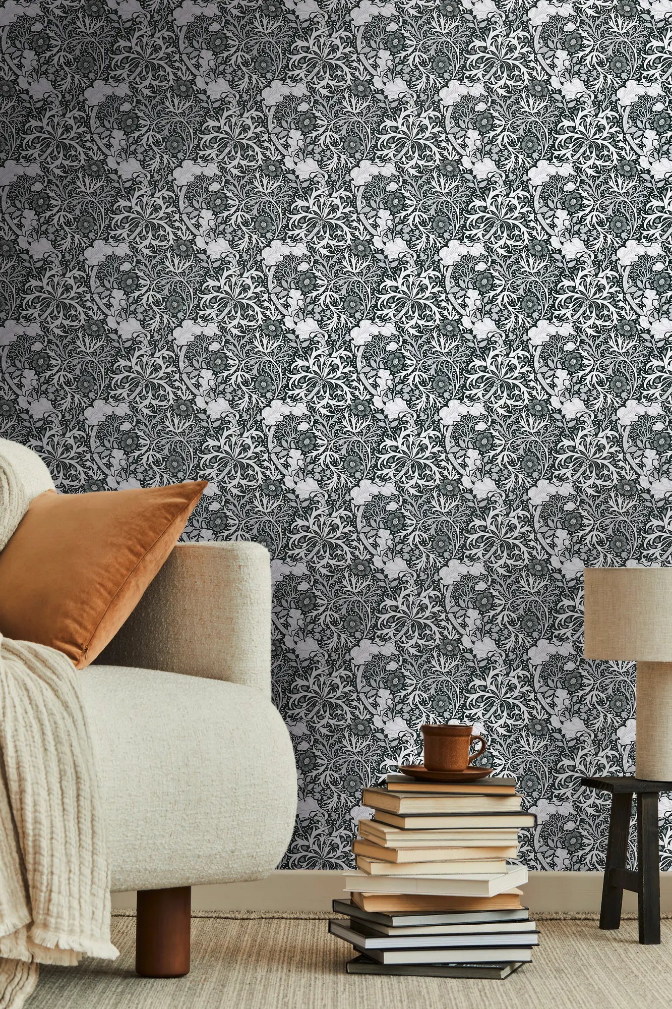 Væg dækket med Symphony of Eve - Charcoal tapet i en moderne indretning med en beige stol, brun pude og stabel af bøger, der fremhæver tapetets elegante design.